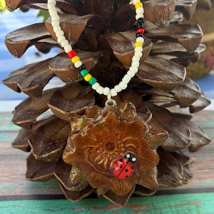 May include: A necklace featuring a brown, flower-shaped pendant made from a pinecone scale, adorned with a small red and black ladybug figurine. The necklace chain consists of white seed beads interspersed with small red, yellow, and green beads.