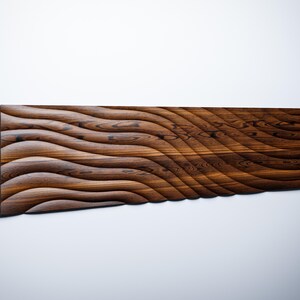 Op de afbeelding: Een lang, rechthoekig houten wandpaneel met een golvende, geribbelde textuur. Het paneel is gemaakt van donkerbruin hout met een natuurlijke houtnerf.