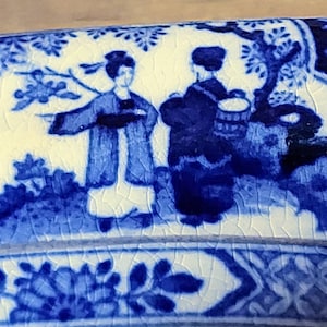 May include: A section of a blue and white ceramic piece with a detailed illustration of two figures in traditional dress. The design includes floral and tree motifs, with a cracked glaze effect.