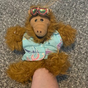 May include: A plush toy of the character Alf, wearing sunglasses and a blue Hawaiian shirt with a floral pattern. The toy has brown fur and a tan snout. The sunglasses have a red and green lens.