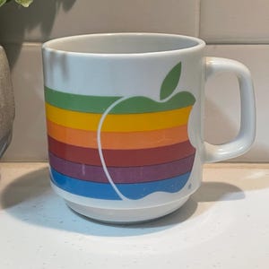 May include: White ceramic mug featuring the iconic Apple logo in a rainbow design. The mug has a rounded base and a sturdy handle. The Apple logo is a prominent graphic element, adding a touch of retro style to this drinkware.