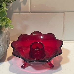 May include: A ruby red glass candle holder with a flower-shaped design. The candle holder has a central cup for the candle and rests on three small feet. The background includes a white tiled wall and a plant in a gray pot.