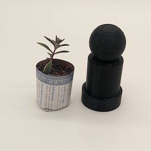 May include: A small potted plant in a newspaper wrap next to a black 3D-printed chess piece. The plant has green and purple leaves. The chess piece is a pawn with a spherical top and cylindrical base.