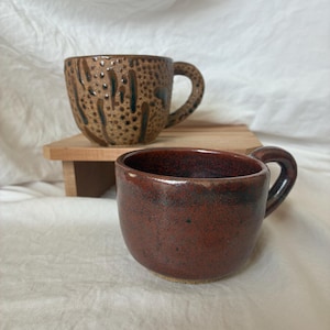 May include: Two ceramic mugs. One is brown with a speckled pattern and dark brown drips. The other is a solid reddish-brown color. Both mugs have a rounded shape and a handle. They are sitting on a small wooden stand.
