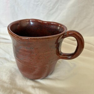 May include: A handmade ceramic mug with a glossy, deep reddish-brown glaze and a speckled texture. The mug features an irregular, organic shape with a wide, rounded rim and a thick, curved handle attached to the side of the vessel.