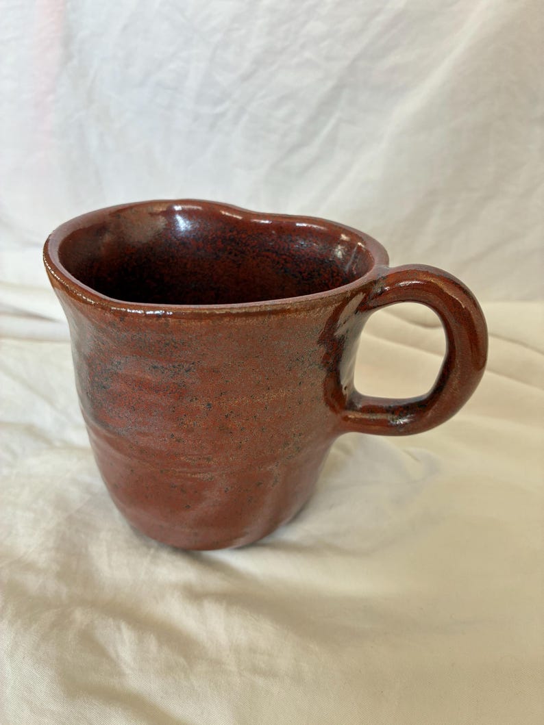 May include: A handmade ceramic mug with a glossy, deep reddish-brown glaze and a speckled texture. The mug features an irregular, organic shape with a wide, rounded rim and a thick, curved handle attached to the side of the vessel.
