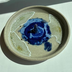 Op de afbeelding: Een handgemaakte keramische ringhouder. De ringhouder is een rond schaaltje met een verhoogd midden. Het schaaltje is beige met blauwe en witte accenten. Het midden is diepblauw.