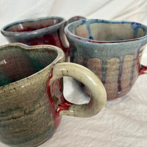 May include: Three handmade ceramic mugs with irregular, organic shapes and thick, curved handles. The mugs feature reactive glazes in shades of deep red, earthy brown, and muted blue, with visible drip patterns and textured, speckled surfaces.