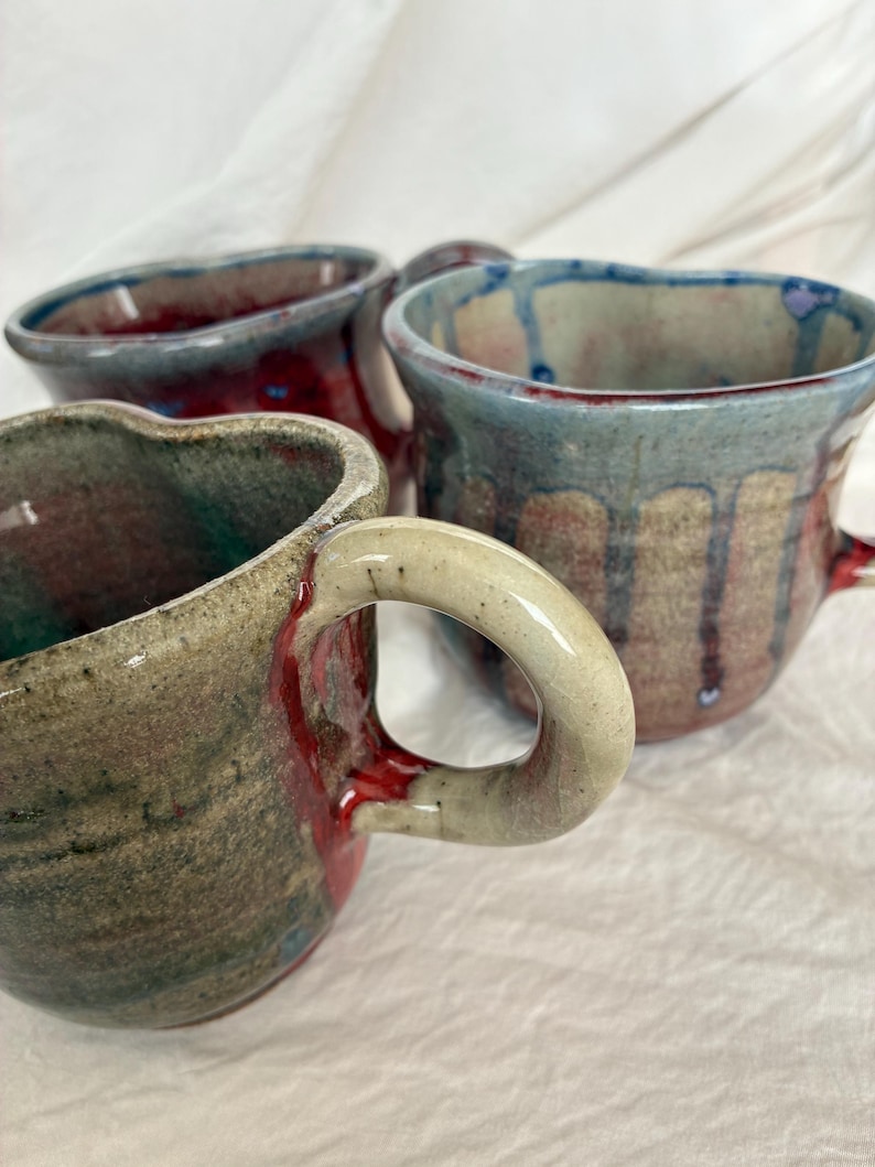 May include: Three handmade ceramic mugs with irregular, organic shapes and thick, curved handles. The mugs feature reactive glazes in shades of deep red, earthy brown, and muted blue, with visible drip patterns and textured, speckled surfaces.