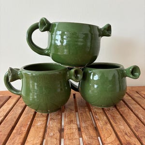 Puede incluir: Tres tazas de cerámica verde hechas a mano apiladas sobre una superficie de madera. Cada taza tiene un asa única y dos pequeñas protuberancias en los lados. Las tazas son de color verde vibrante y parecen estar esmaltadas.