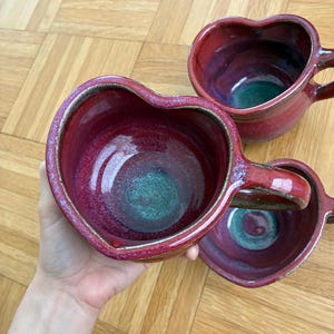 Op de afbeelding: Een witte keramische mok met een zwarte handgreep en de tekst "But First, Coffee" in een vetgedrukte, zwarte letter. De mok staat tegen een vage achtergrond, wat een gezellige setting suggereert.