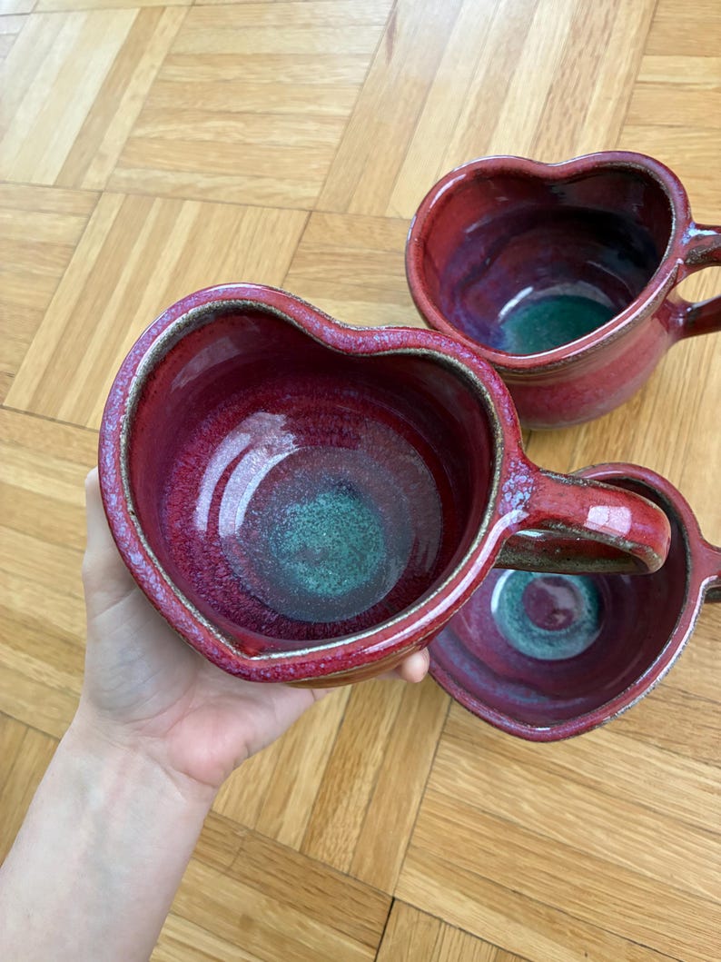 May include: Three heart-shaped ceramic mugs featuring a glossy red and magenta exterior glaze with a contrasting teal green interior base. Each mug has a sturdy handle and a textured, speckled rim, showcasing a handcrafted pottery aesthetic.