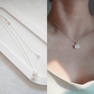 May include: A delicate silver chain necklace with a small white flower pendant. The necklace is shown on a white surface and on a person's neck.