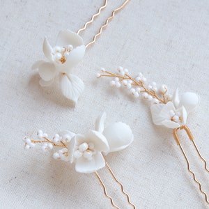 Puede incluir: Tres horquillas para el cabello con flores blancas y tallos de alambre dorado. Cada horquilla presenta delicadas flores esculpidas y peque&ntilde;as cuentas perladas. Las horquillas est&aacute;n dispuestas sobre una superficie clara y texturizada.