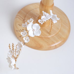Puede incluir: Un tocado de novia con flores de porcelana blanca y perlas de imitación sobre una estructura de alambre dorado. El diseño floral incluye grandes flores abiertas y capullos más pequeños, creando una estética elegante. Ideal para una boda o evento especial.