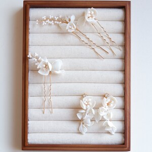 Puede incluir: Una colecci&oacute;n de accesorios para el cabello y pendientes florales blancos exhibidos en una bandeja de madera. El conjunto incluye varias horquillas para el cabello con delicados detalles de flores y cuentas, y un par de pendientes colgantes.