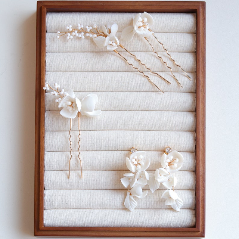 Puede incluir: Una colecci&oacute;n de accesorios para el cabello y pendientes florales blancos exhibidos en una bandeja de madera. El conjunto incluye varias horquillas para el cabello con delicados detalles de flores y cuentas, y un par de pendientes colgantes.