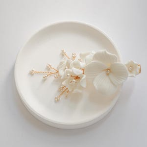 Puede incluir: Un peine de pelo floral blanco con detalles de perlas y alambre dorado. El peine está en un plato blanco.