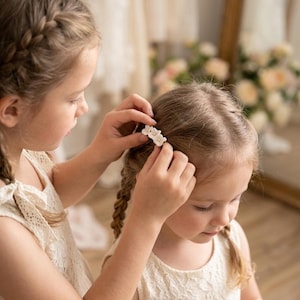 Puede incluir: Una pequeña pinza para el cabello con tres flores blancas y diminutos centros dorados. El accesorio floral está colocado en cabello castaño claro, mostrando un diseño delicado con pétalos dispuestos en un grupo para sujetar el pelo.