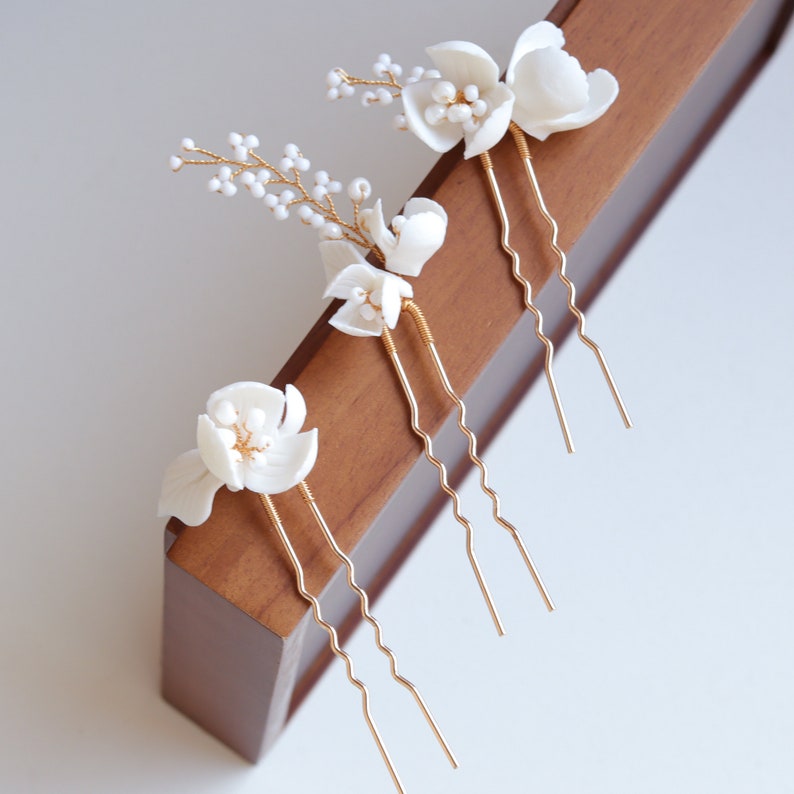 Puede incluir: Cuatro horquillas florales blancas con tallos y detalles dorados. Las horquillas presentan delicados dise&ntilde;os florales tridimensionales y peque&ntilde;as ramitas con cuentas. Las horquillas est&aacute;n dispuestas sobre una superficie de madera, lo que sugiere un tema nupcial o de ocasi&oacute;n especial.