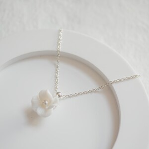 May include: A delicate silver chain necklace with a white flower pendant. The flower has a pearl in the center.