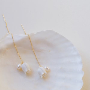 May include: A pair of delicate gold chain earrings with white flower charms and pearl accents. The earrings are displayed on a white seashell.
