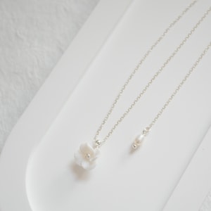 May include: A delicate silver chain necklace with a white flower pendant and a single pearl charm. The necklace is displayed on a white surface.