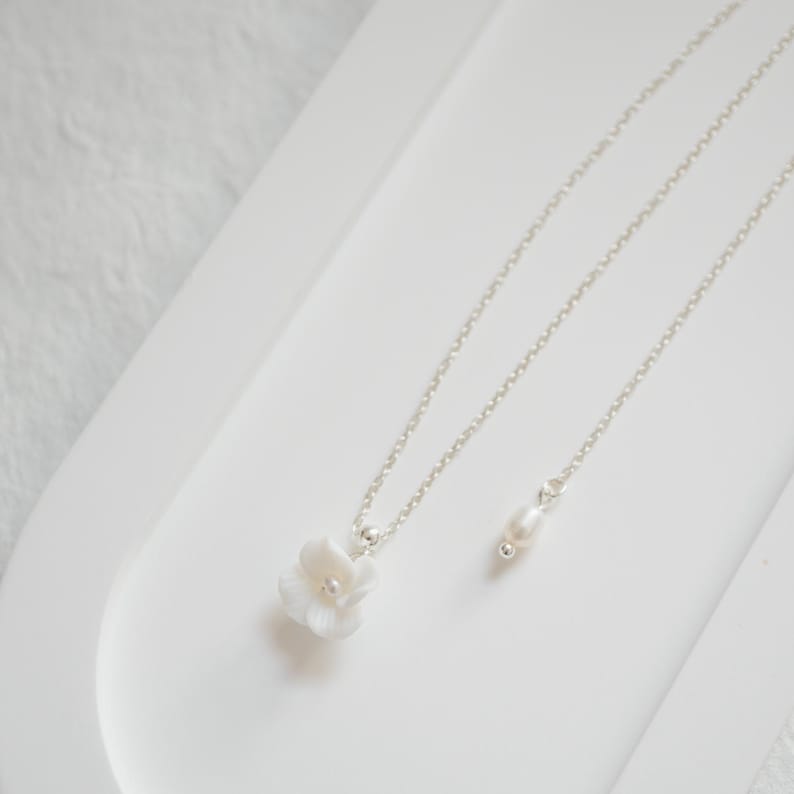 May include: A delicate silver chain necklace with a white flower pendant and a single pearl charm. The necklace is displayed on a white surface.