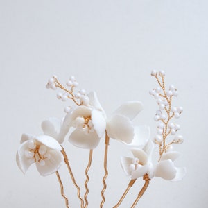 Puede incluir: Primer plano de varios pasadores de pelo florales blancos. Cada pasador presenta una delicada flor blanca con un grupo de peque&ntilde;as cuentas blancas y un tallo de alambre dorado. Los pasadores est&aacute;n dispuestos sobre un fondo blanco liso.
