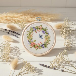 May include: A circular embroidery hoop containing white fabric with a cross-stitched floral wreath in shades of pink, purple, green, and yellow. A small blue dragonfly is stitched in the centre of the wreath. The hoop has a silver metal hanger.