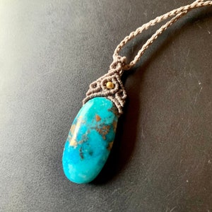 Puede incluir: Un colgante de piedra turquesa con detalles dorados, colgado de un collar de cordón marrón.