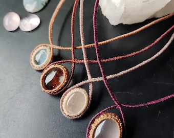 May include: Five macrame necklaces with oval gemstones in shades of blue, brown, and pink. The necklaces have cords in complementary colors. Additional gemstones and a quartz crystal are also visible.