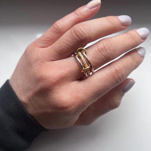 May include: A multi-band ring with a silver band and gold accents. The ring features a row of small, clear stones and is worn on a person's finger. The ring has a modern, geometric design.