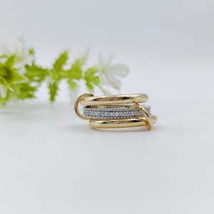 Könnte beinhalten: Ein goldfarbener Ring mit mehreren Bändern, von denen eines mit kleinen, klaren Edelsteinen besetzt ist. Die Bänder sind ineinander verschlungen und erzeugen einen geschichteten Effekt. Der Ring befindet sich vor einem weißen Hintergrund mit einem Hauch von Grün.