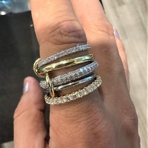 Pode incluir: Um anel de várias bandas com bandas douradas e prateadas, algumas incrustadas com pequenas pedras brilhantes. O anel é usado num dedo, mostrando o seu design intrincado e várias texturas. As bandas são empilhadas, criando uma peça ousada e atraente.
