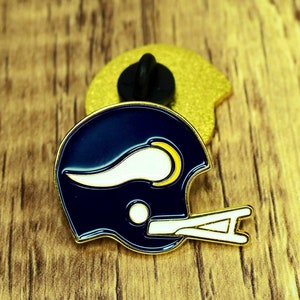 May include: A gold and blue enamel pin featuring a football helmet with a white horn. The pin is on a wooden surface.