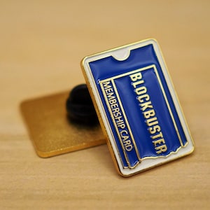 May include: Gold-coloured pin with a blue and white design. The pin features the text "BLOCKBUSTER MEMBERSHIP CARD".