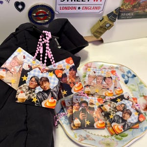 May include: Several square photo charms with images of people, cupcakes, and stars, linked by a pink beaded chain. The charms are displayed on a floral plate. A black garment is in the background.