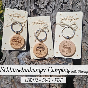 Peut inclure: Trois porte-clés en bois avec des motifs de camping. Chaque porte-clés comprend un pendentif rond en bois avec une scène de camping, attaché à un anneau en métal et à une petite carte en bois. Le texte "Eine Kleinigkeit" est imprimé sur les cartes. Le bas de l'image contient le texte "Schlüsselanhänger Camping inkl. Displays".