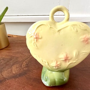 May include: A yellow heart-shaped ceramic vase with a green base and pink floral details. The vase has a handle on the top.