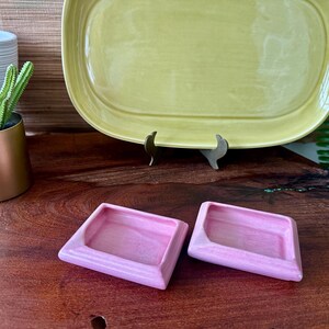 May include: Two small, pink, square-shaped ceramic dishes with a raised rim sit on a wooden surface. A large, oval, yellow-green ceramic platter is in the background, supported by a metal stand. A small gold pot with a cactus is on the left.