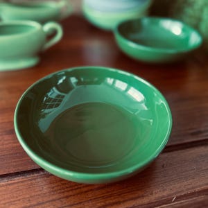 May include: A collection of green ceramic dishware, including bowls and a cup with a handle. The glossy finish reflects light, highlighting the smooth surfaces. The items are arranged on a dark wooden table, creating a contrast.
