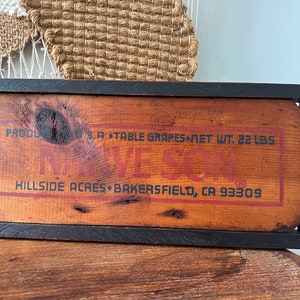 May include: A rectangular wooden sign with a black frame. The sign has a reddish-brown background with the text "U.S.A. TABLE GRAPES NET WT. 22 LBS" and "NAVE SON" in red. The sign also includes "HILLSIDE ACRES BAKERSFIELD, CA 93309".