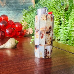 May include: A rectangular lighter featuring a mosaic exterior composed of polished, translucent stone tiles in shades of cream, beige, and dark brown. The top mechanism is metallic silver with a small red ignition button on the side.