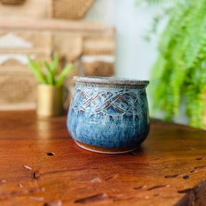 May include: A handcrafted ceramic pot with a blue glaze and a textured, woven-like pattern. The pot has a brown rim and base, and it sits on a wooden surface. The pot is a decorative item.