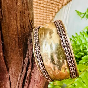 May include: A wide, hammered brass cuff bracelet with silver and copper accents. The bracelet has a textured surface and a braided rope design along the edges. The background includes wood, woven material, and green foliage.