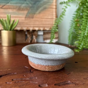 May include: A small, handmade ceramic bowl with a light gray interior and a textured, brown base. The bowl is round with a slightly raised rim. The bowl is sitting on a wooden surface.