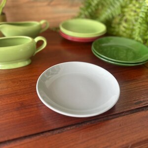 May include: A collection of ceramic tableware, including a light gray plate, green saucers, and a green gravy boat. The items are arranged on a dark wooden surface, with greenery in the background.