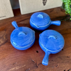 May include: Three small, round, cobalt blue ceramic dishes with lids and handles. The dishes are arranged on a dark wooden surface. The lids have a small, curved handle. The dishes are likely for serving individual portions.