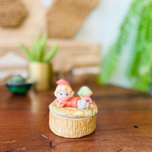 May include: A ceramic trinket box featuring a whimsical elf figurine. The elf, dressed in a red outfit and hat, is lying on a bed of straw. The box has a textured, wood-like appearance. The overall design is charming and decorative.